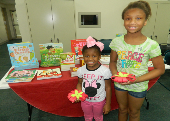 two girls with tomatoes in front of table of books