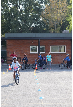 Smiling fifth-grade student at Washington STEM Elementary School riding a bicycle practicing the bike skills drills, while her classmates wait their turn to execute the course. Several adults are observing the students.