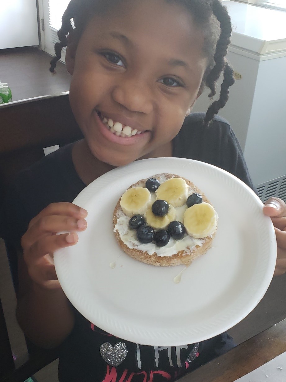 a smiling girl holds up a plate with a bagel decorated with fruit