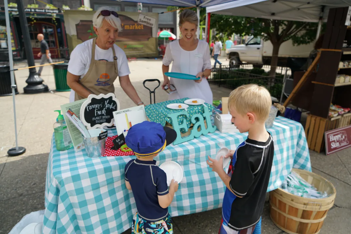 two boys try food from a sample table at a Farmers Market