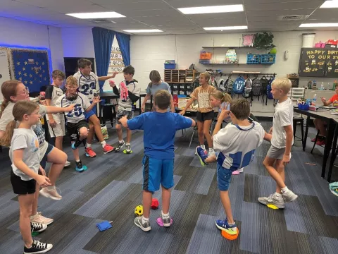 Group of excited 5th graders in althletic clothes in a classroom, standing in a circle, ciking foam dice across the circle to eachother.  