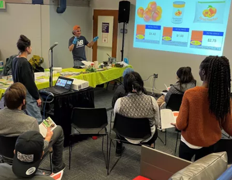 College-aged young adults viewing a presentation led by an instructor compating prices of canned, fresh, and frozen fruit.