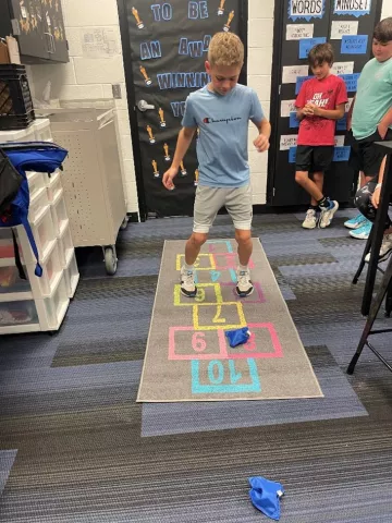 A 5th grade boy plays hopschotch on an indoor mat in his classroom while 2 more boys look on and wait their turn behind him. 